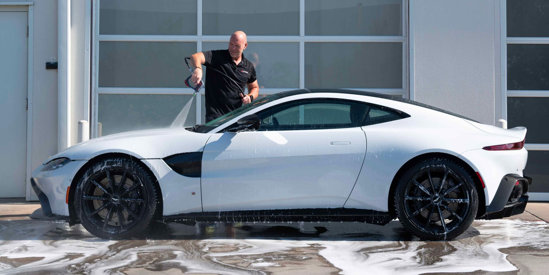 A man washes a white sports car with black wheels in front of a garage on a sunny day.