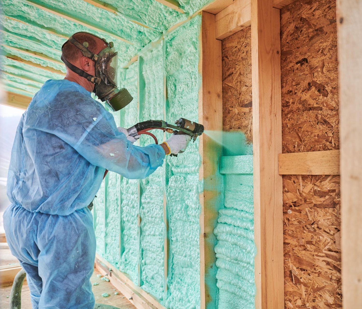 A man wearing a gas mask is spraying insulation on a wall.