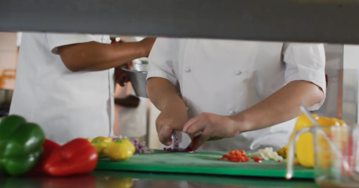 Two chefs are cutting vegetables on a cutting board in a kitchen.