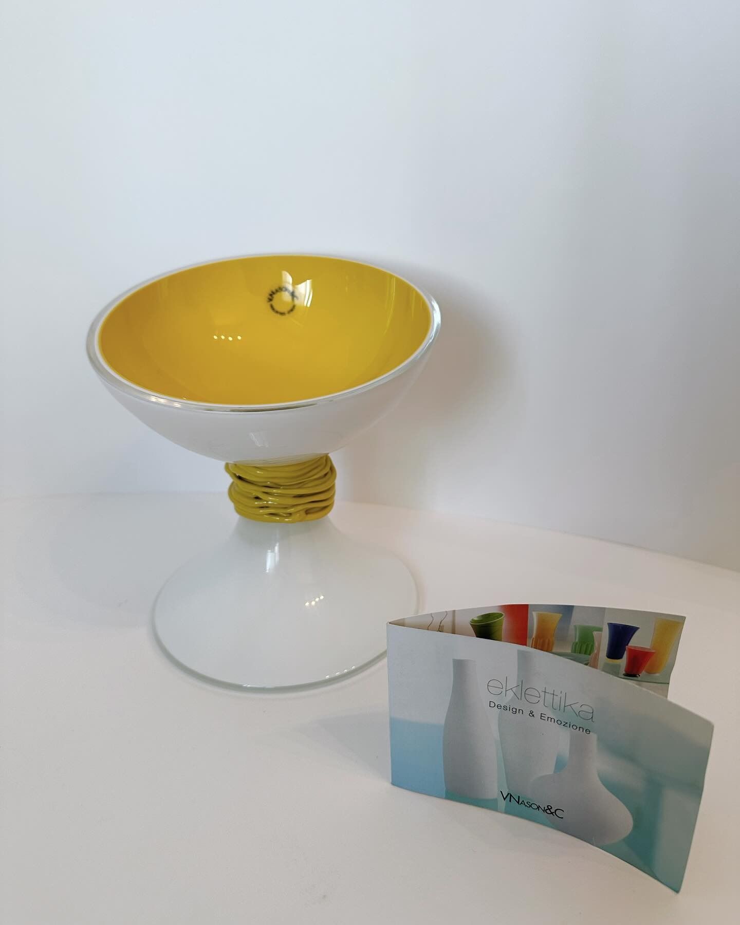 Ciotola decorativa bianca e gialla su piedistallo, con bordo dorato ed etichetta dorata.