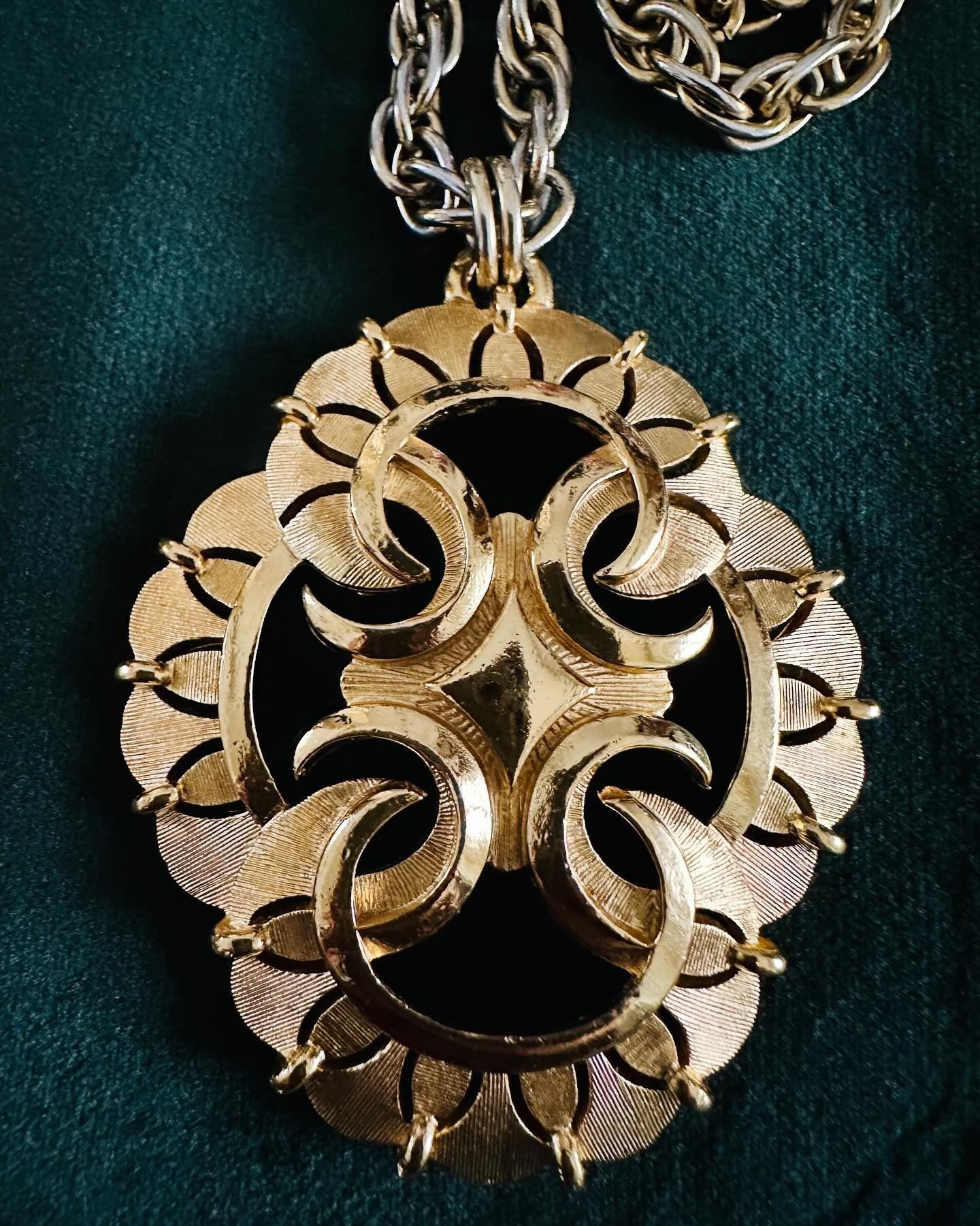 Collana con pendente decorato color oro, con montatura ovale e intricato motivo floreale su sfondo verde scuro.