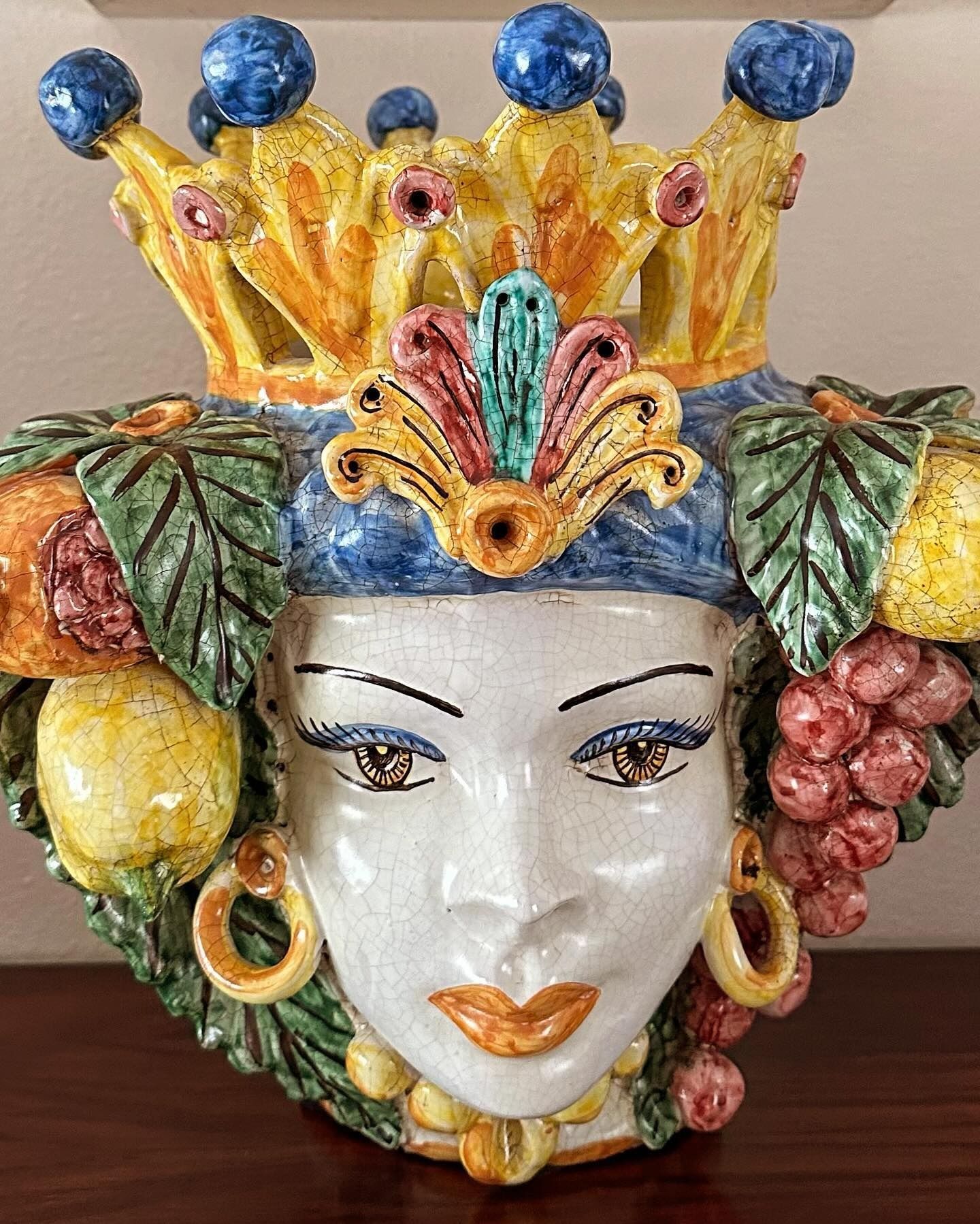 Testa in ceramica con corona e decoro di frutta, viso con capelli blu, orecchini in oro, frutta ai lati, colori vivaci.