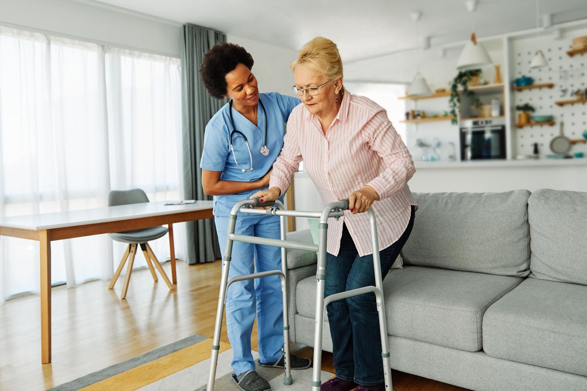 A nurse is helping an elderly woman use a walker in a living room.