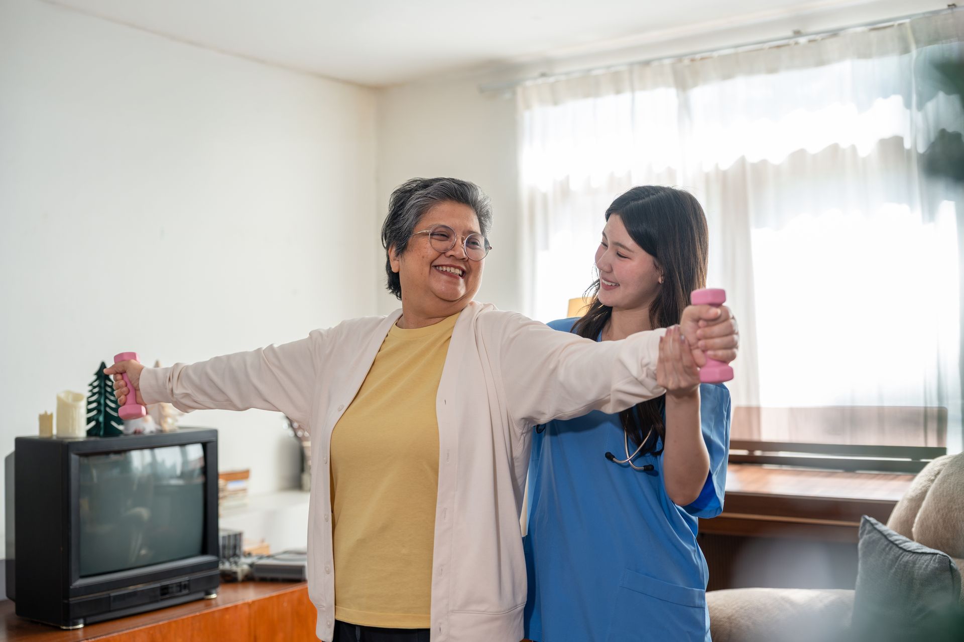 Two women smiling and dancing in a bright living room beside a TV and window