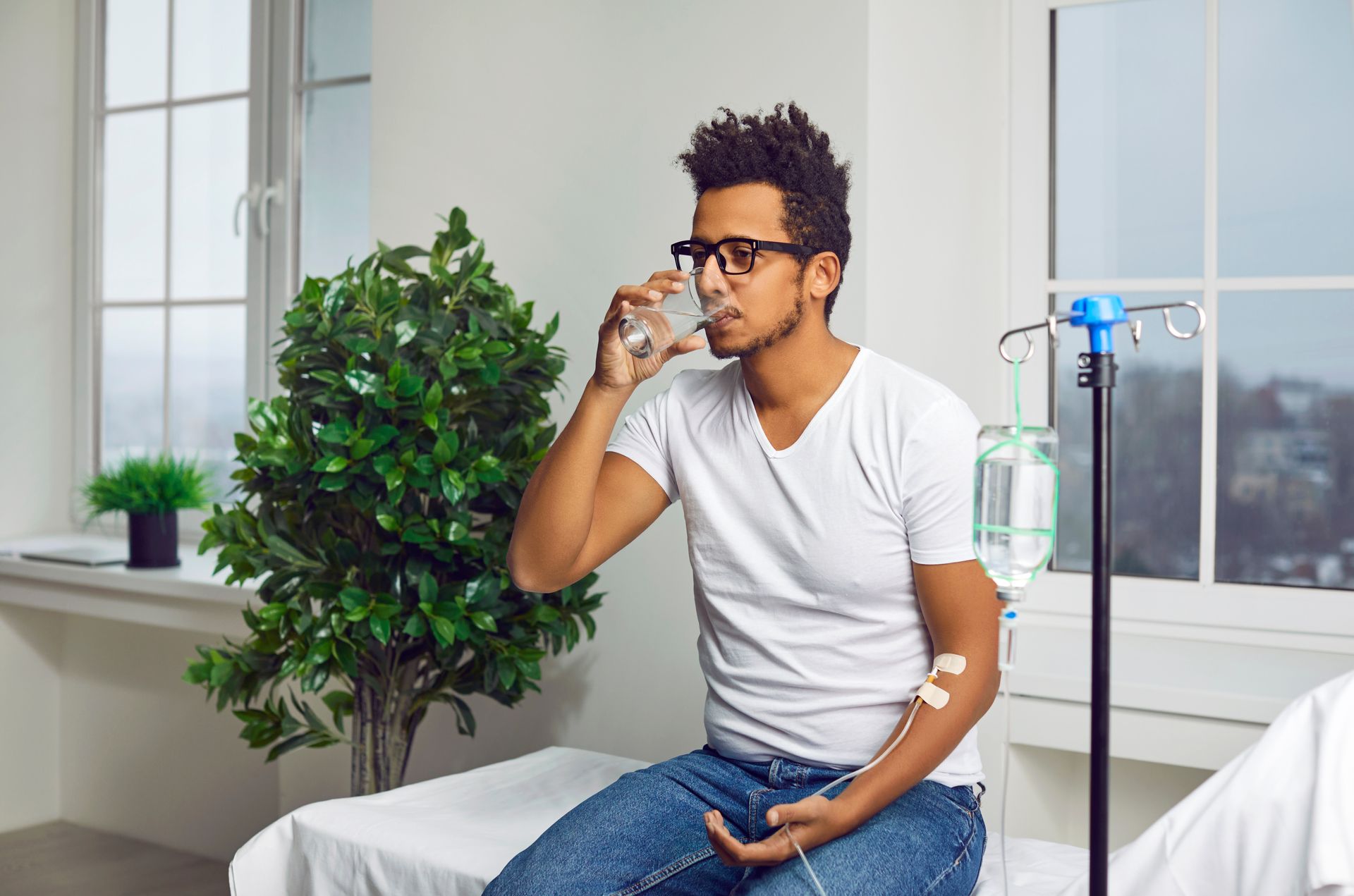 Person drinking from a glass while seated in a bright room with a plant and IV stand nearby
