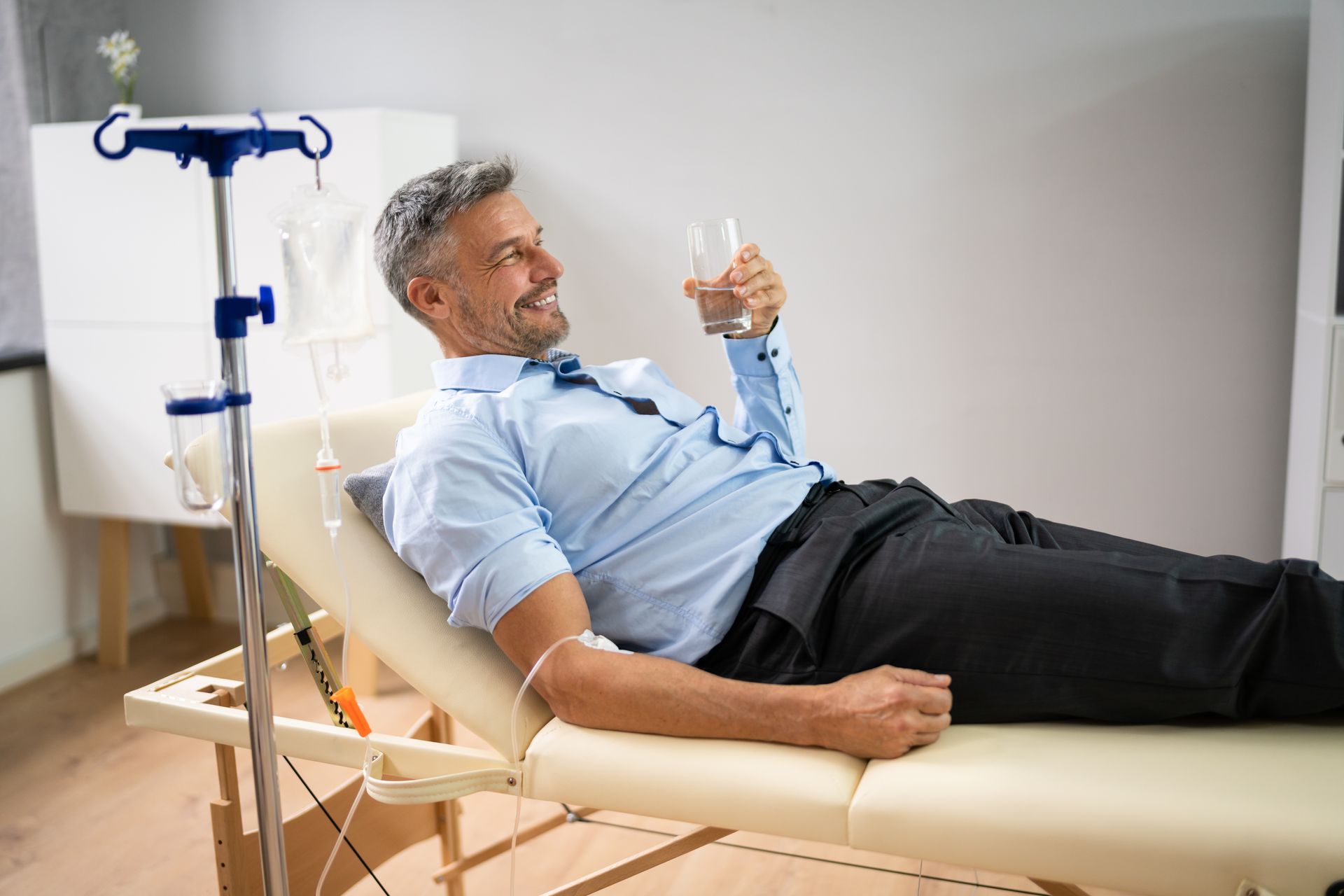 Man reclining on a treatment bed, holding a phone, with an IV stand nearby.