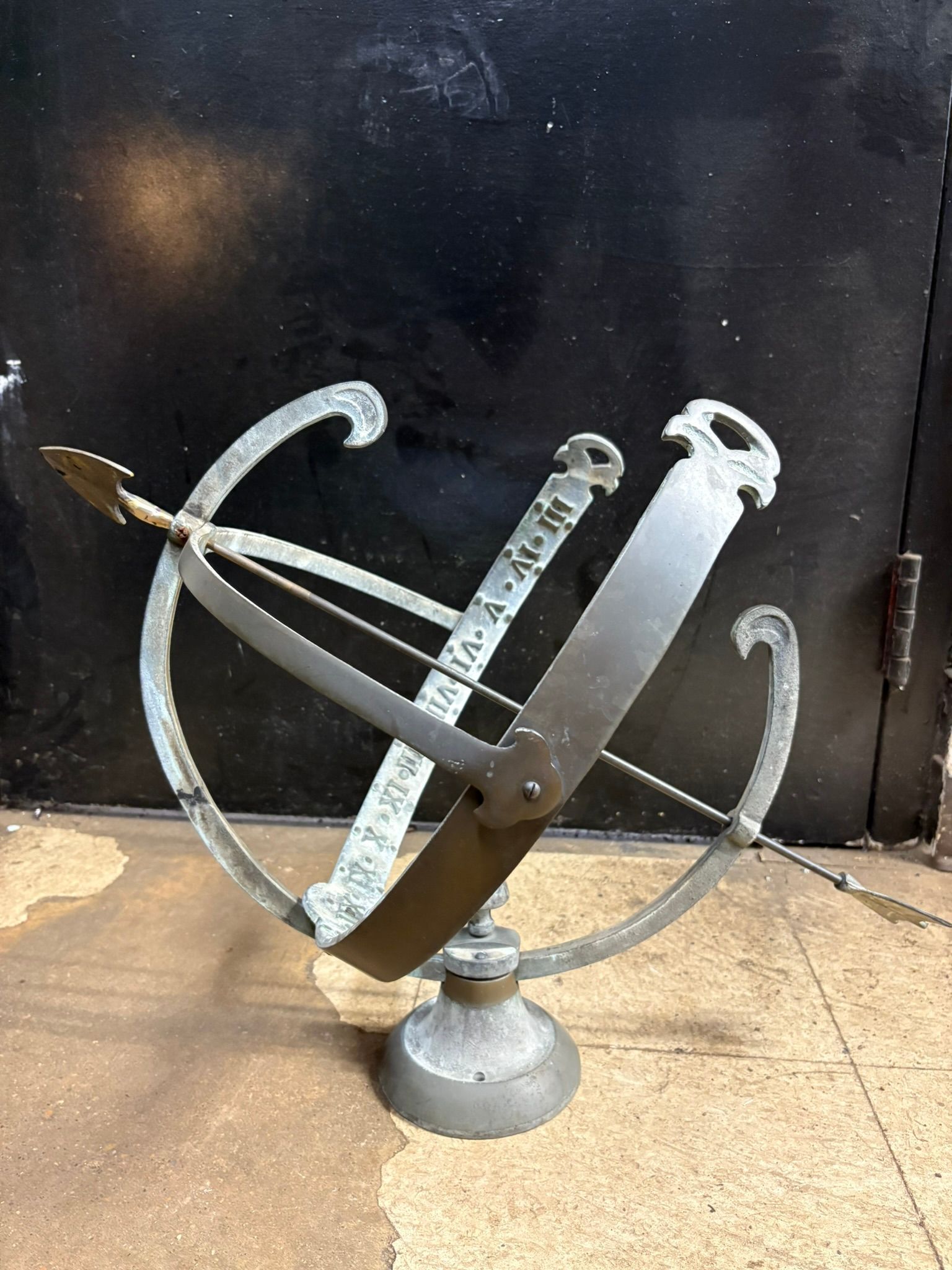 Metal armillary sundial sculpture on a pedestal against a dark wall outdoors.