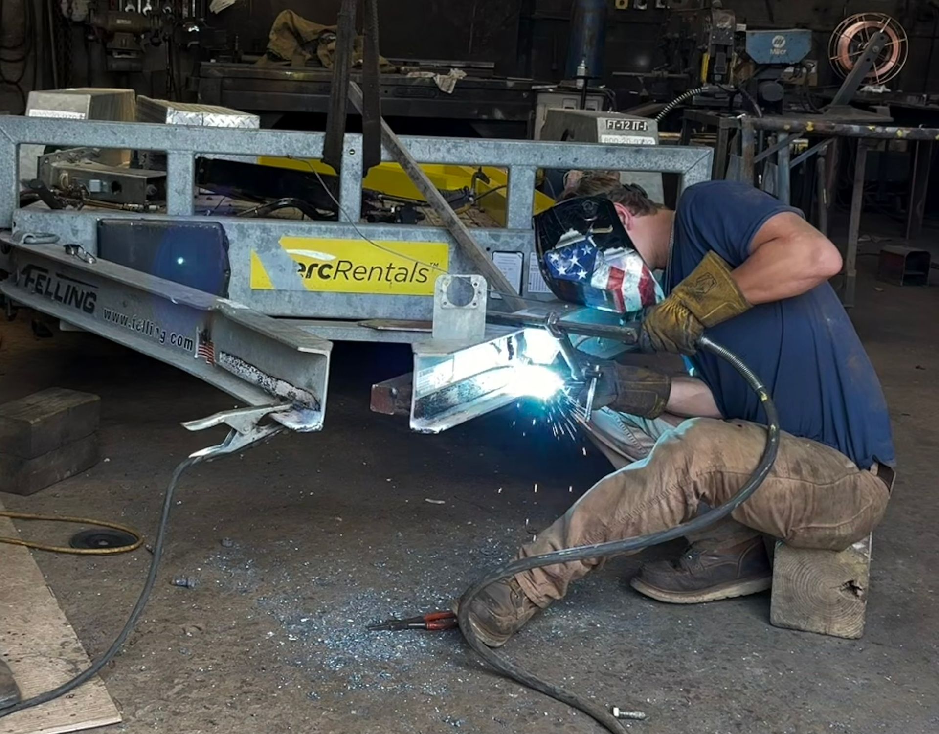 Worker welding under a trailer in a garage, bright sparks flying.