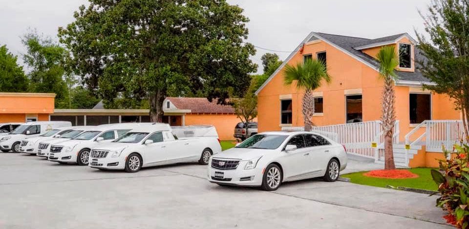 White hearses and cars parked outside an orange building with a green lawn and trees.