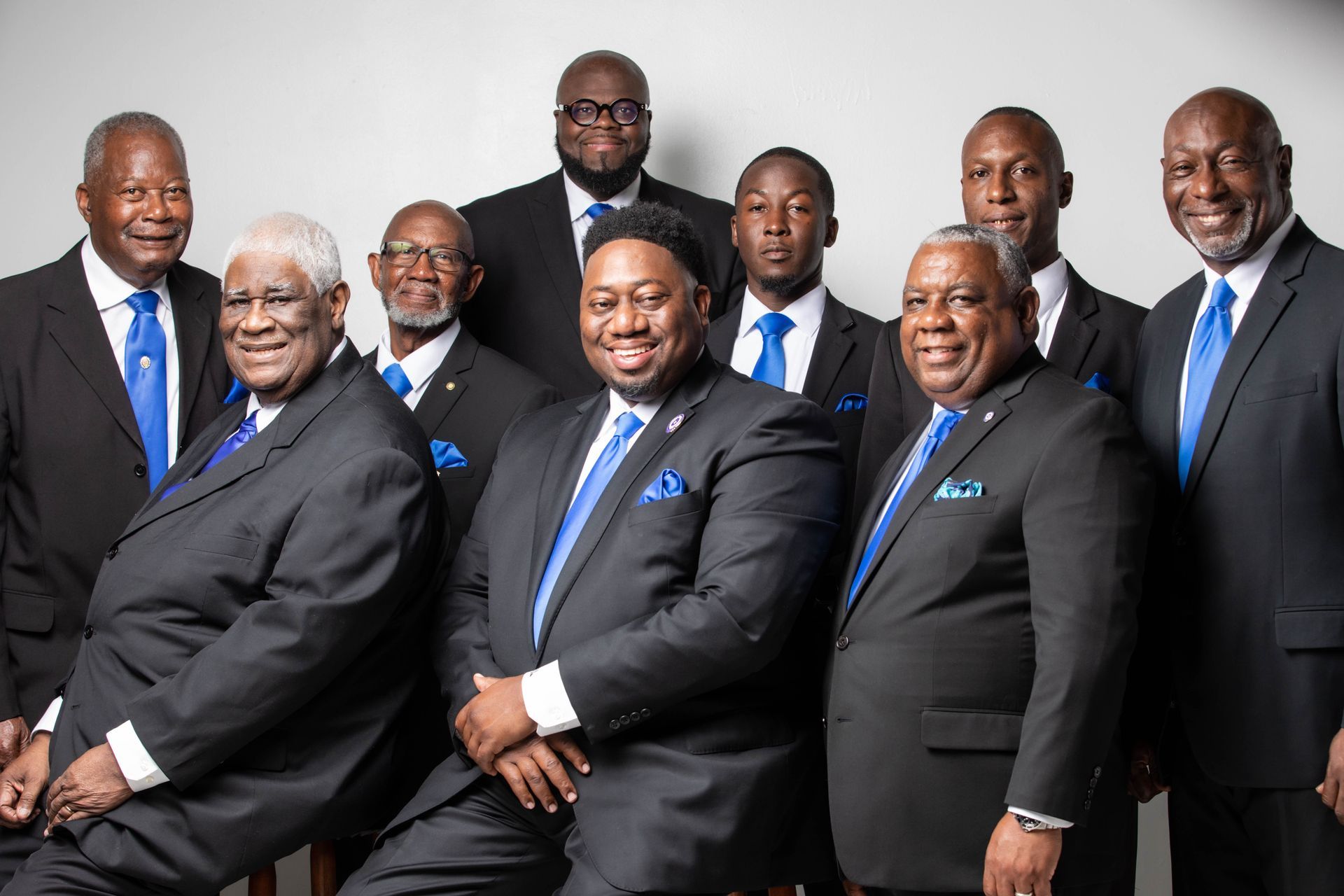 Group of men in suits, some with blue ties and pocket squares, posing indoors against a white background.