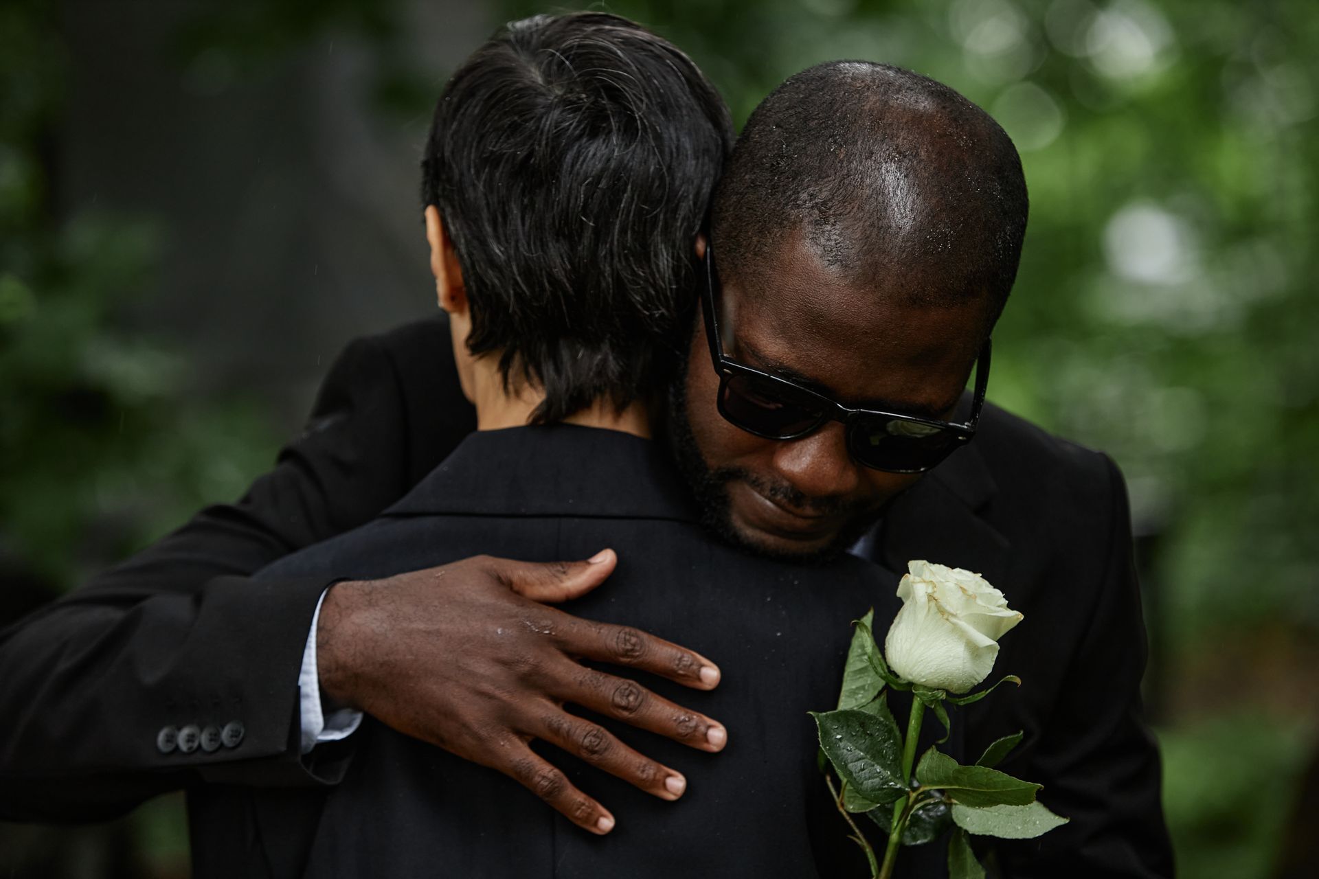 Man in sunglasses hugs another person, holding a white rose, outdoors.