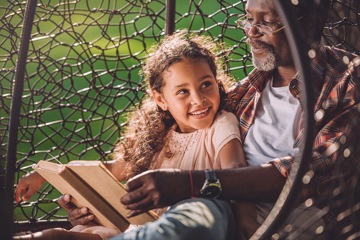 A person and a child reading a book, smiling, in a woven chair outdoors.