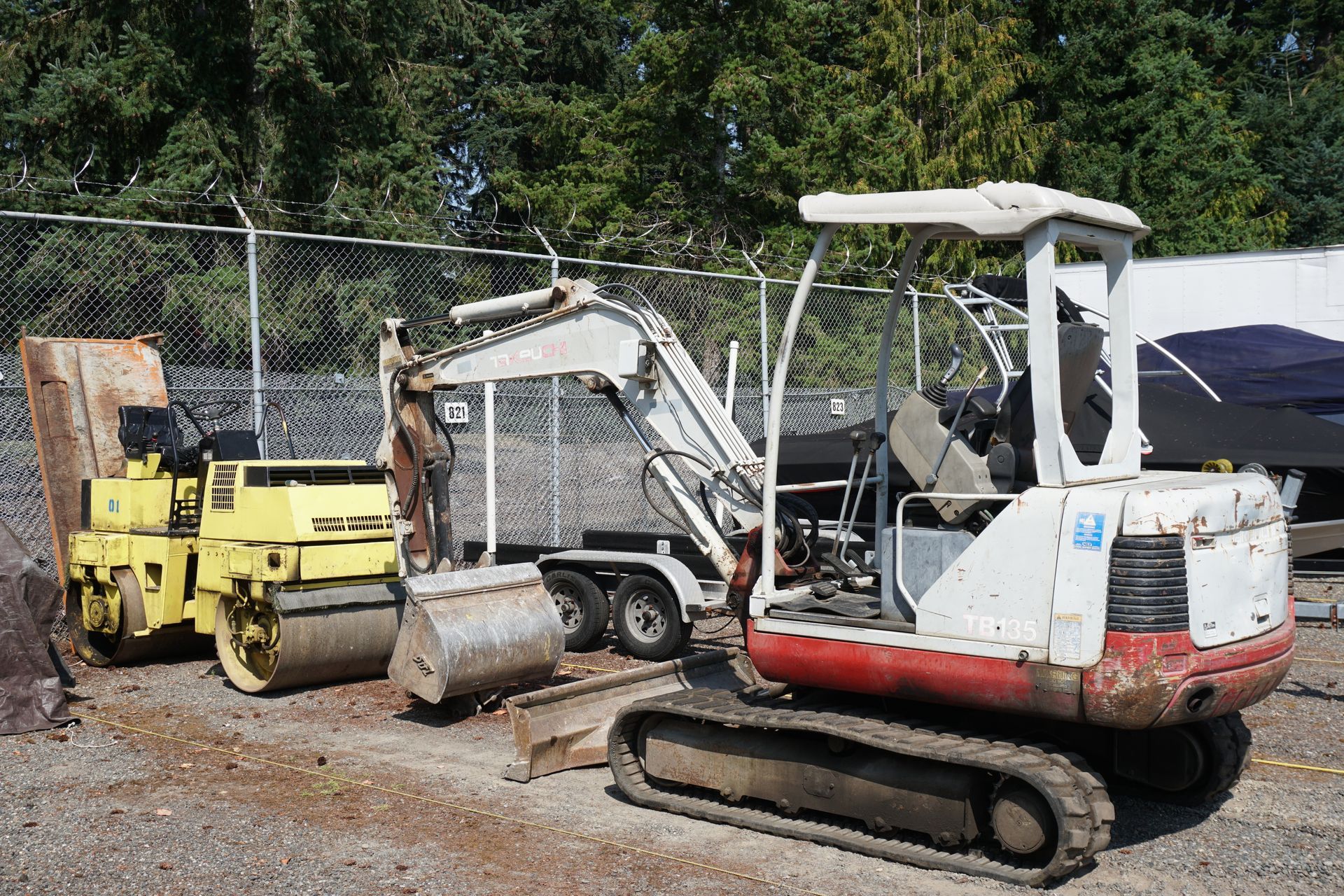 Yellow roller, white excavator, and equipment trailer against a fence.
