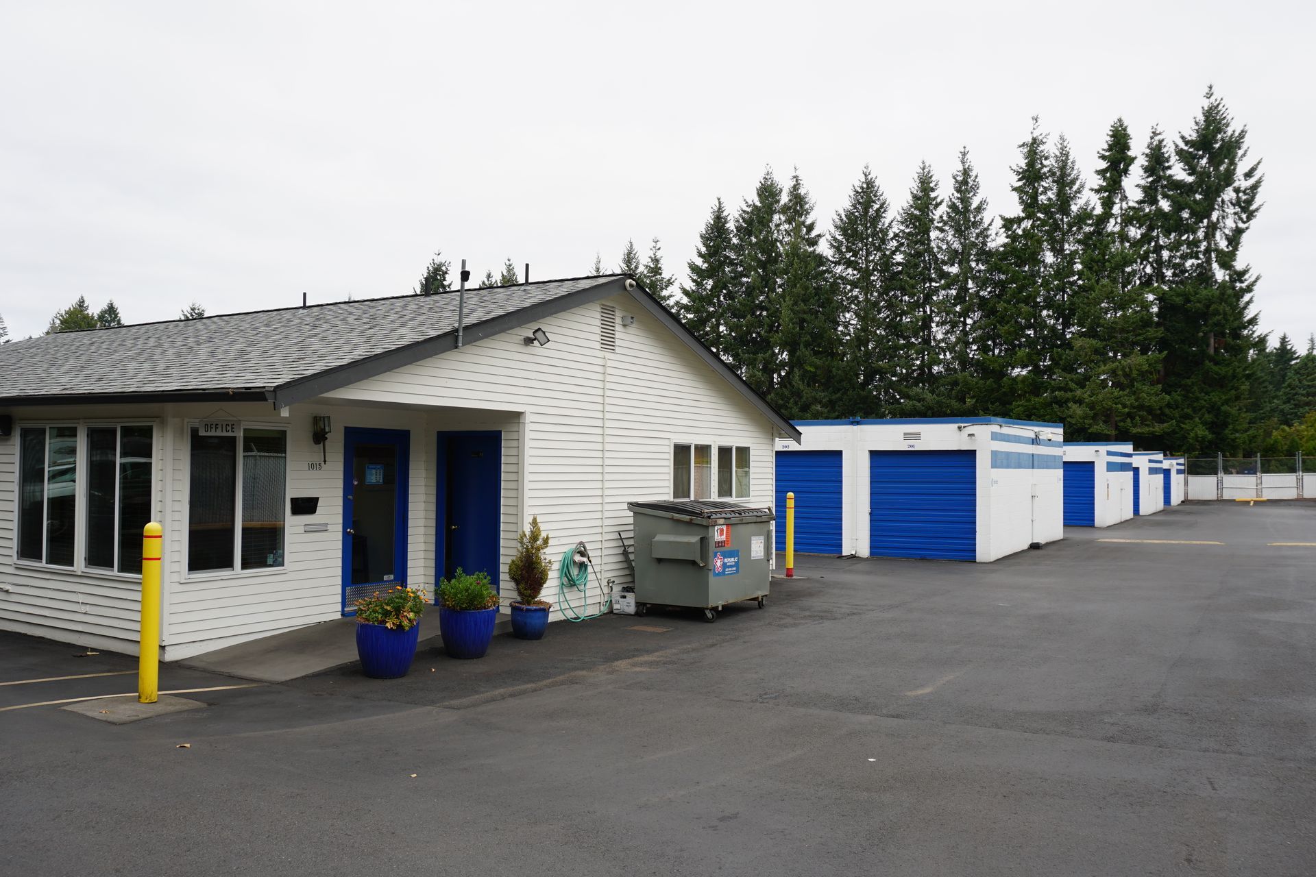 A white storage facility building with blue doors and storage units, a dumpster, and trees in the background.