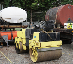 Yellow and black road roller, with dark drum, in a yard with tanks.