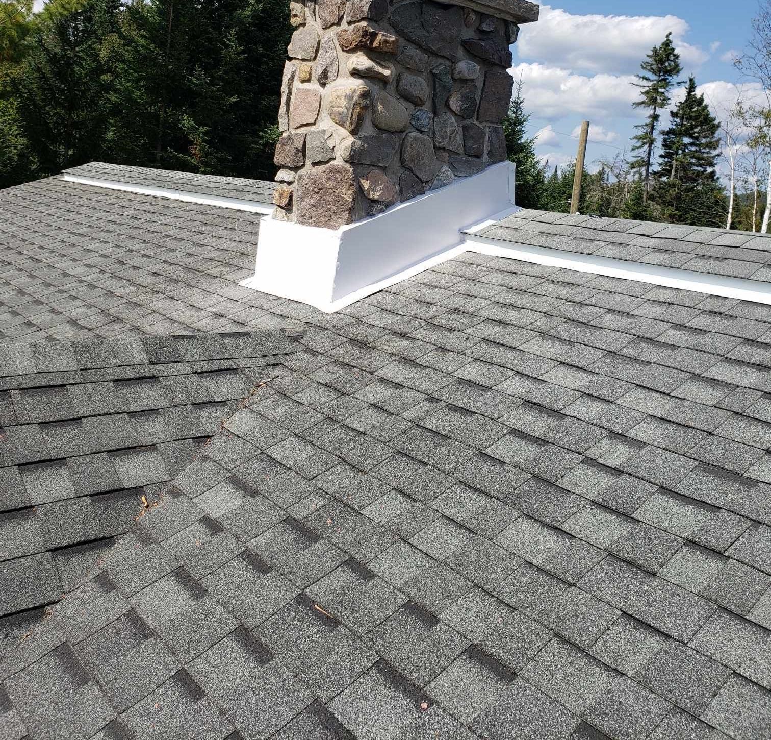 Toit en bardeaux gris avec une cheminée en pierre et des solins blancs sur fond de ciel bleu.
