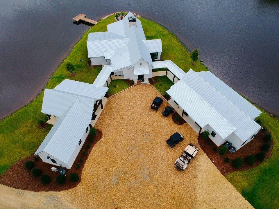 Aerial view of a white modern farmhouse with a metal roof on a grassy point of land, next to a lake, and a gravel driveway.