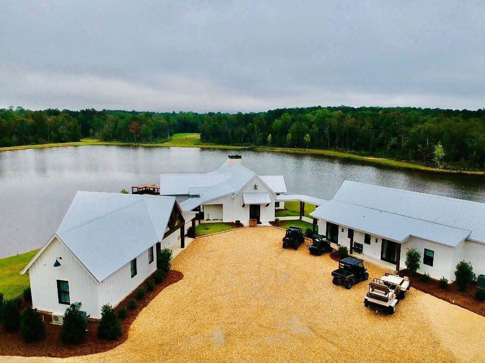 White buildings with metal roofs by a lake, several vehicles on gravel. Trees in background.