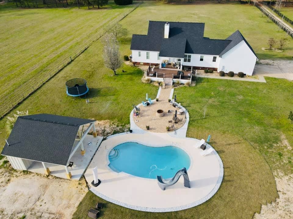 Aerial view: White house with black roof, pool, patio, and cabana on grassy property.