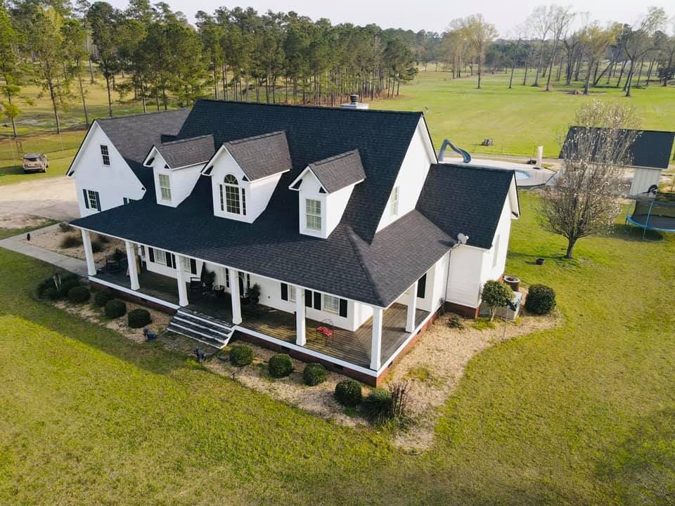 White farmhouse with a black roof and large porch, on a grassy lot.
