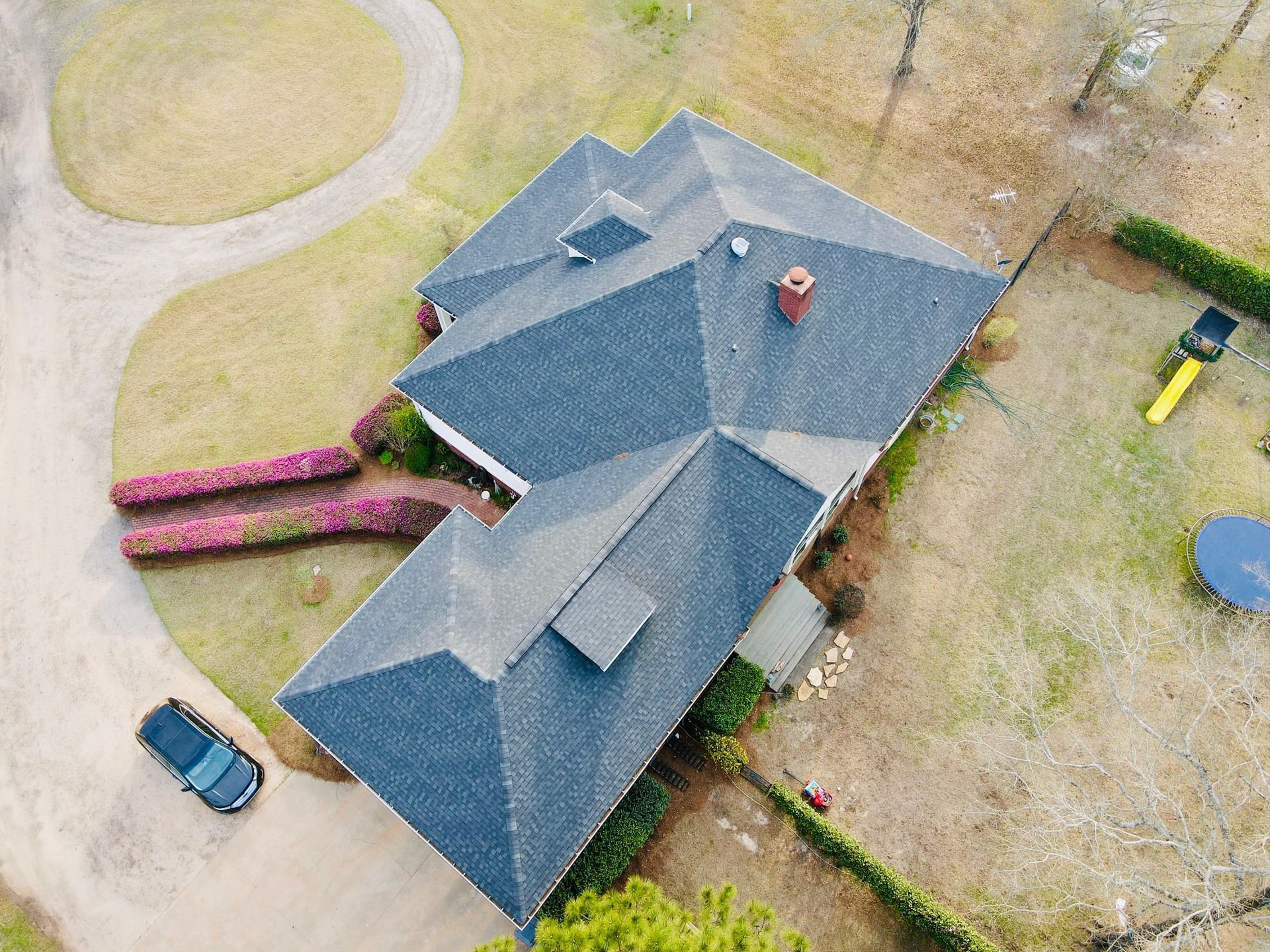 Aerial view of a house with a dark roof, driveway, and surrounding lawn with a parked car.
