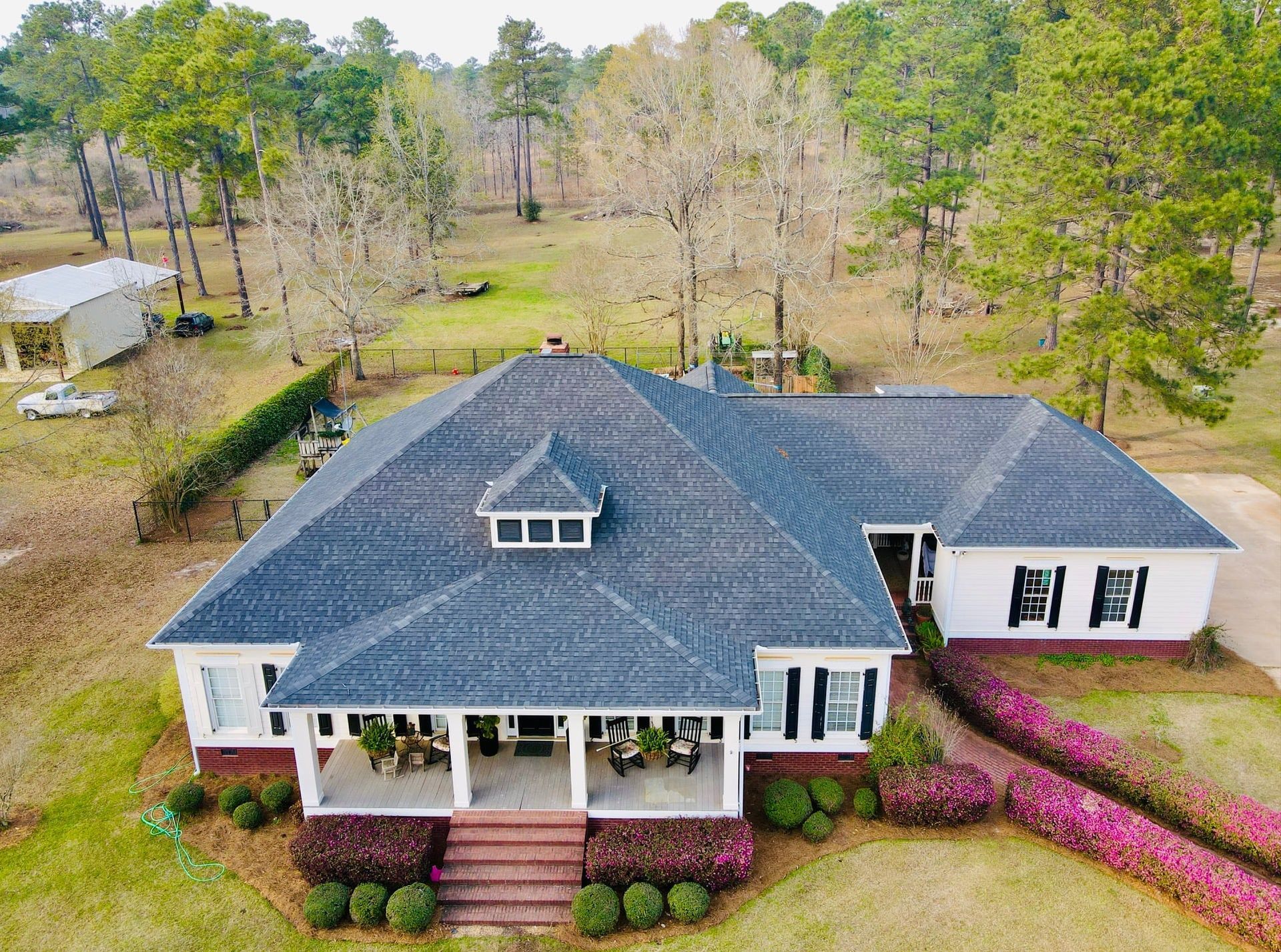 White house with dark gray roof, front porch, and pink flowers in a grassy yard, surrounded by trees.
