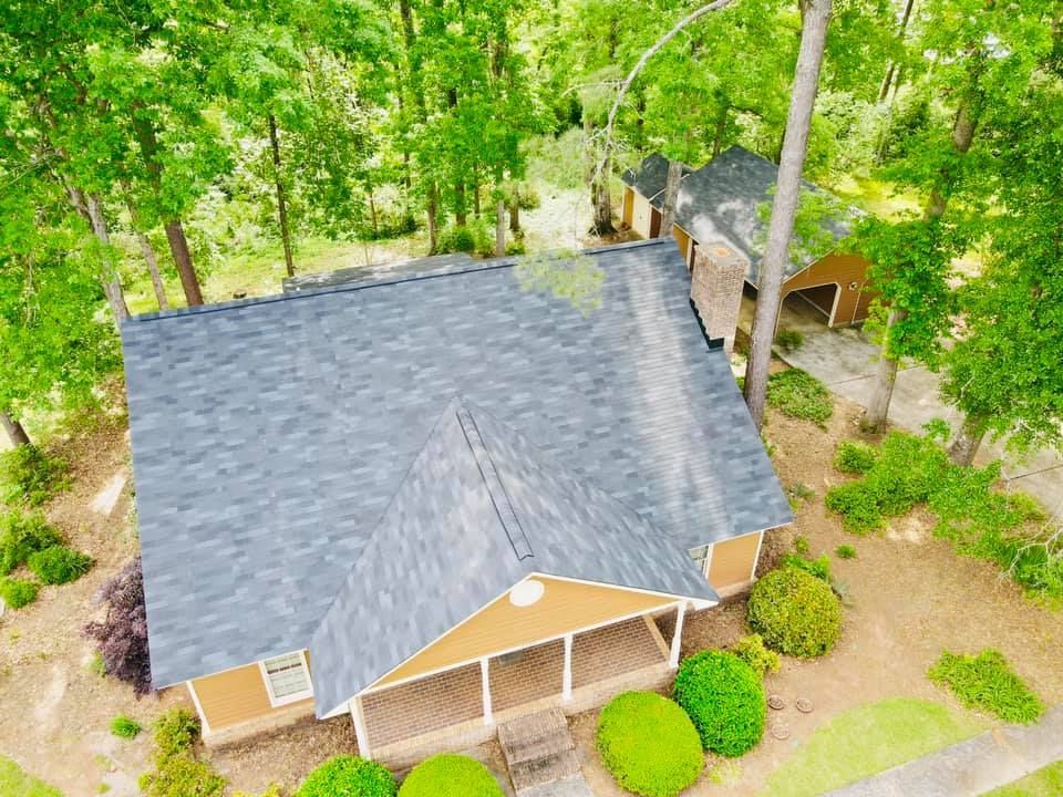 Aerial view of a house with a gray roof and tan siding, surrounded by trees and greenery.