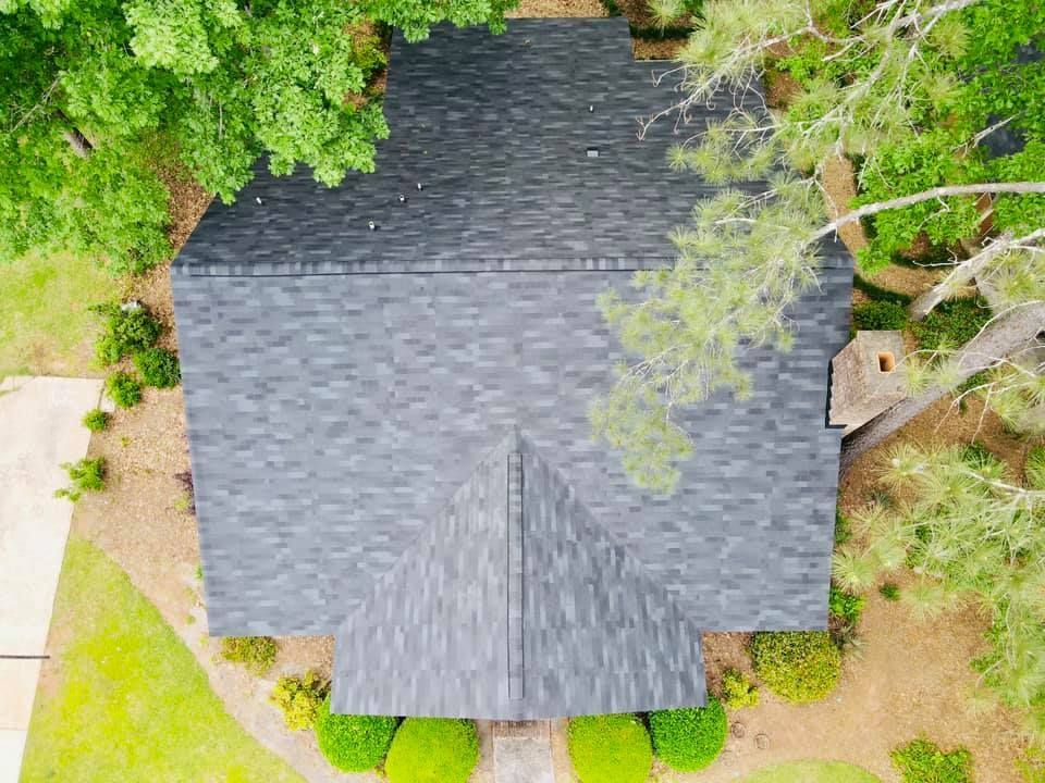 Aerial view of a dark gray asphalt shingle roof surrounded by green trees and bushes, with a path and lawn visible.