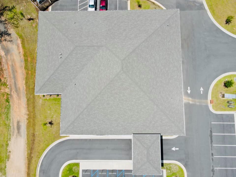 Overhead view of a building with a gray shingle roof, surrounded by parking lot and grassy areas.