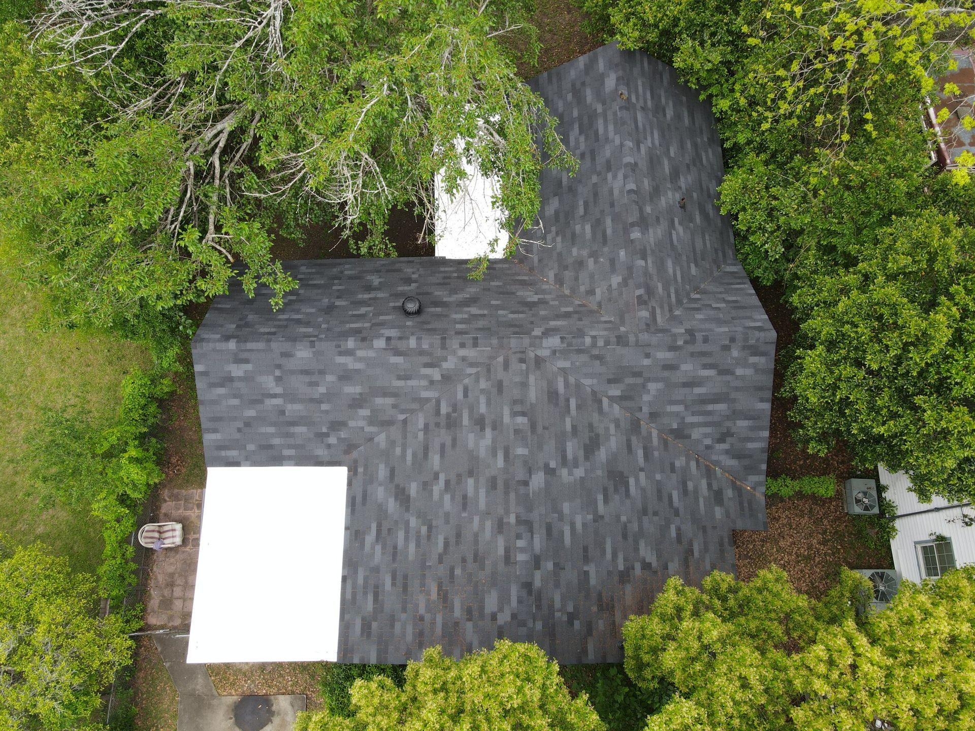 Overhead view of a house with a dark gray shingle roof, surrounded by green trees. A white rectangular surface is visible.