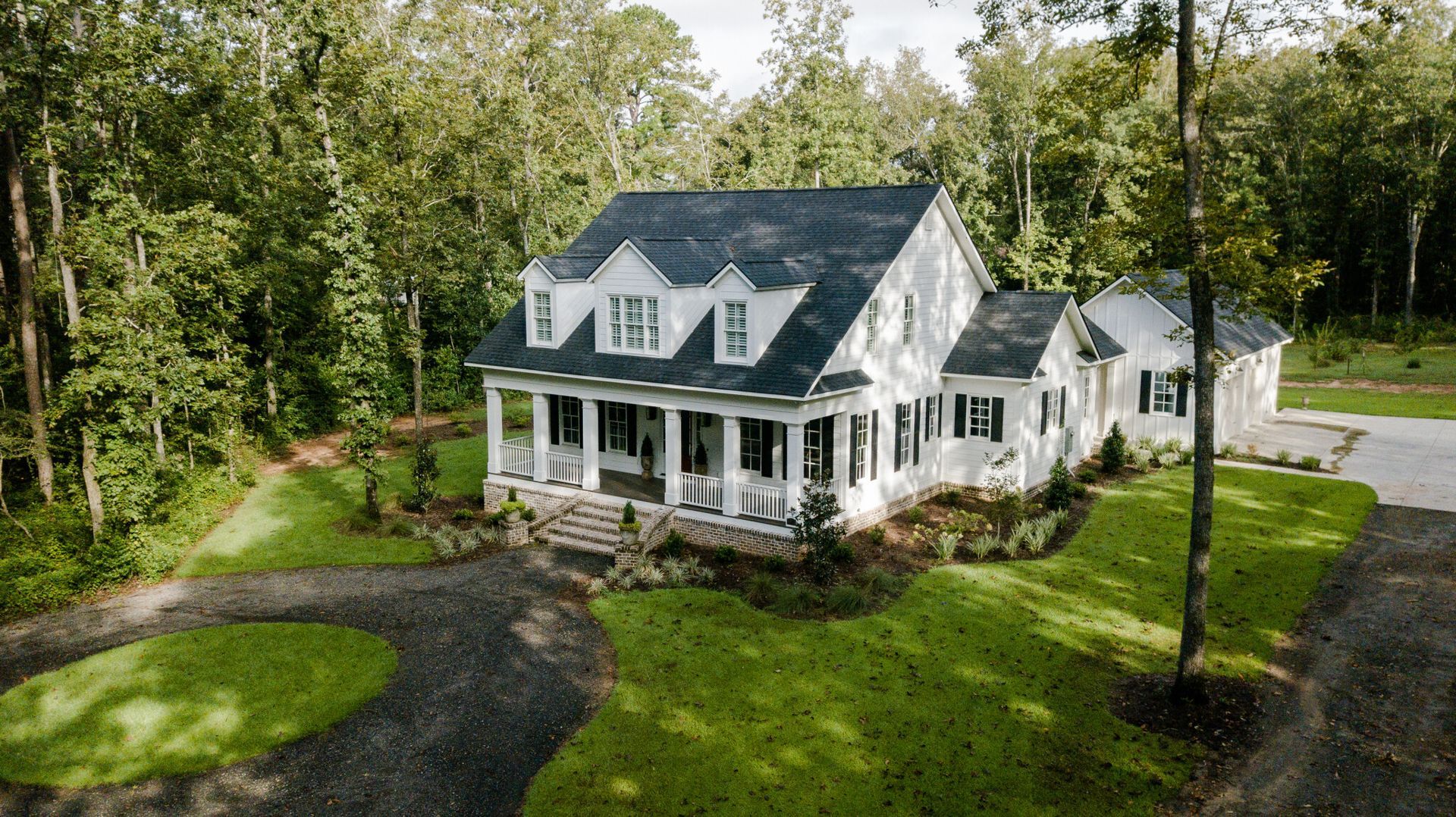 White two-story house with black shutters, porch, and a dark shingle roof, surrounded by green grass and trees.