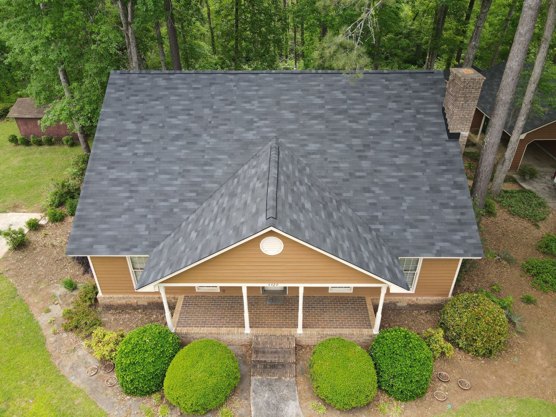Aerial view of a brown house with a dark gray roof surrounded by trees and green bushes.