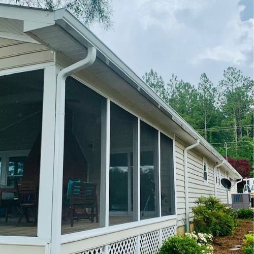 Screened porch with white trim and gutters, attached to a tan house with a backdrop of trees.