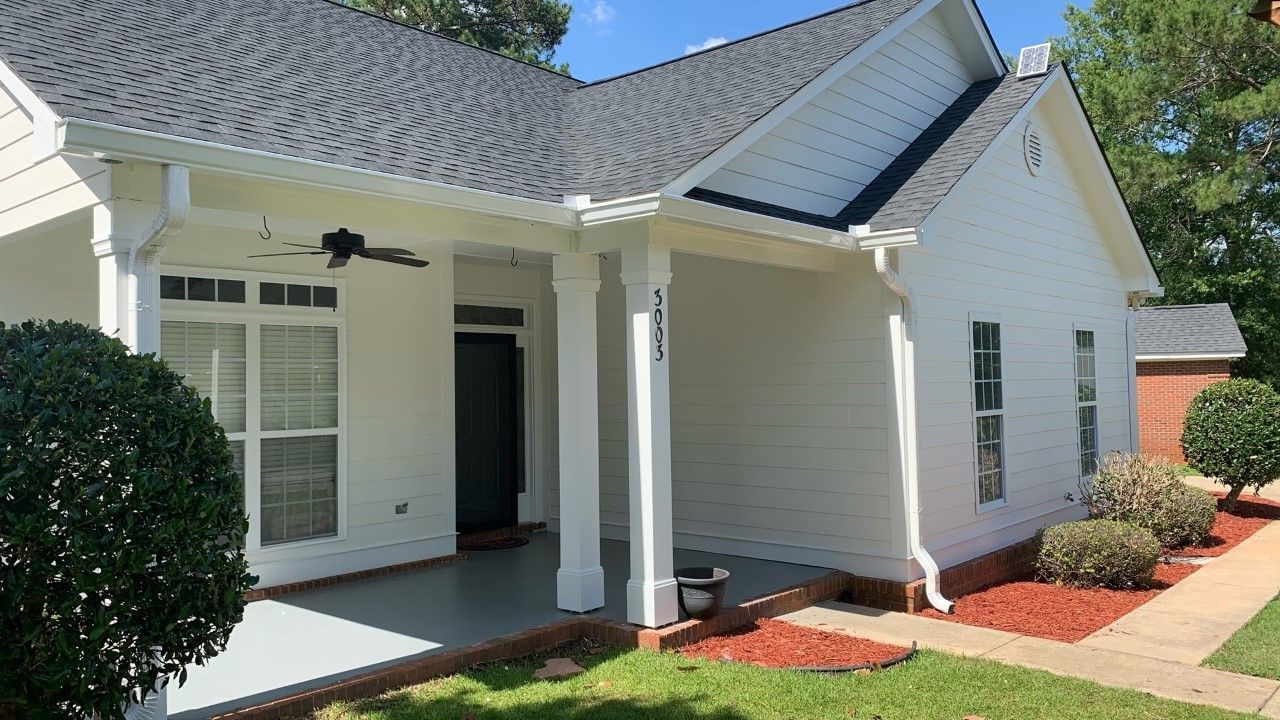 White house with dark roof and porch, gray floor, green bushes, and red mulch.