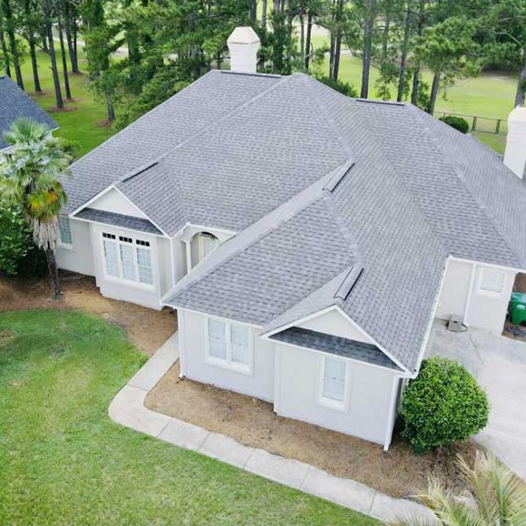 Aerial view of a light gray house with a gray roof and white trim, surrounded by green grass and trees.