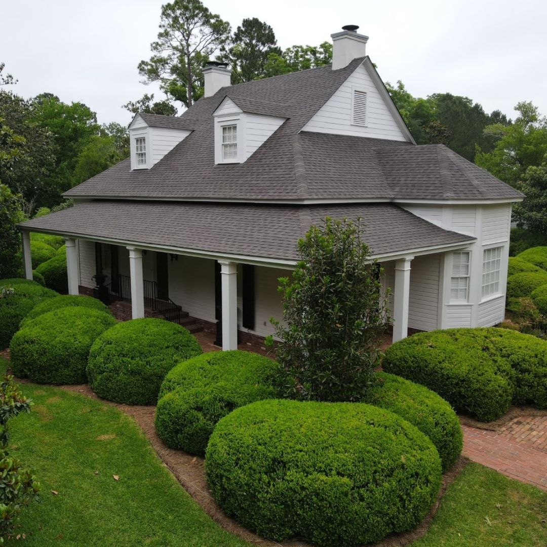 White house with a porch, dormers, and dark roof, surrounded by green shrubs and trees.