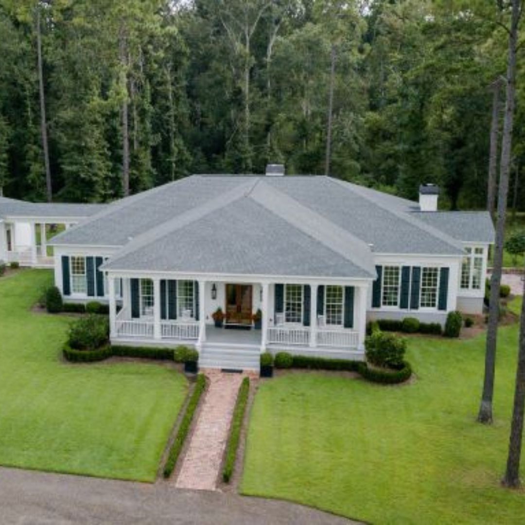 White house with green shutters, gray roof, and brick walkway surrounded by green lawn and trees.
