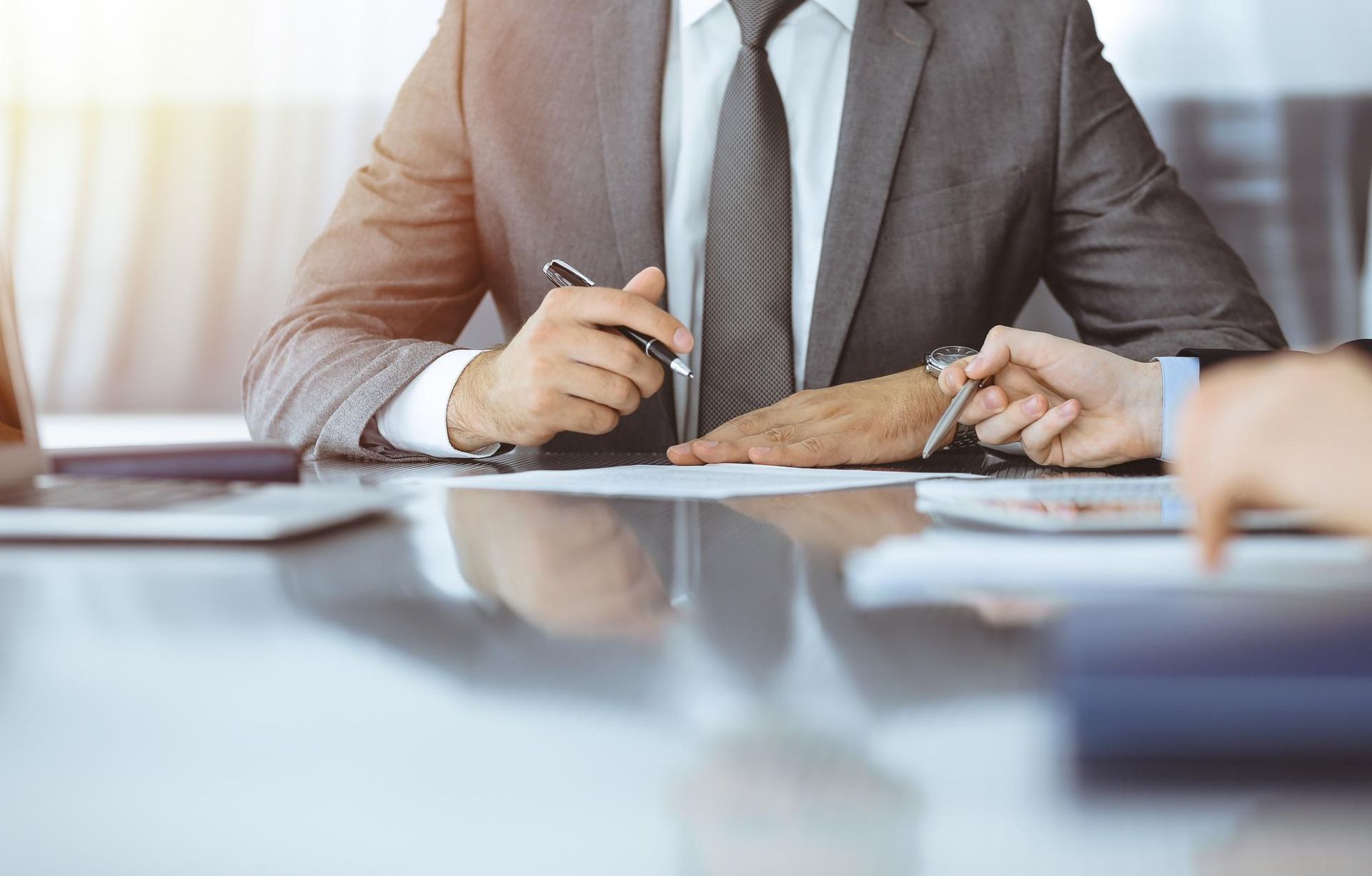Two men in suits at a desk, one signing a document as the other holds a pen, in an office.