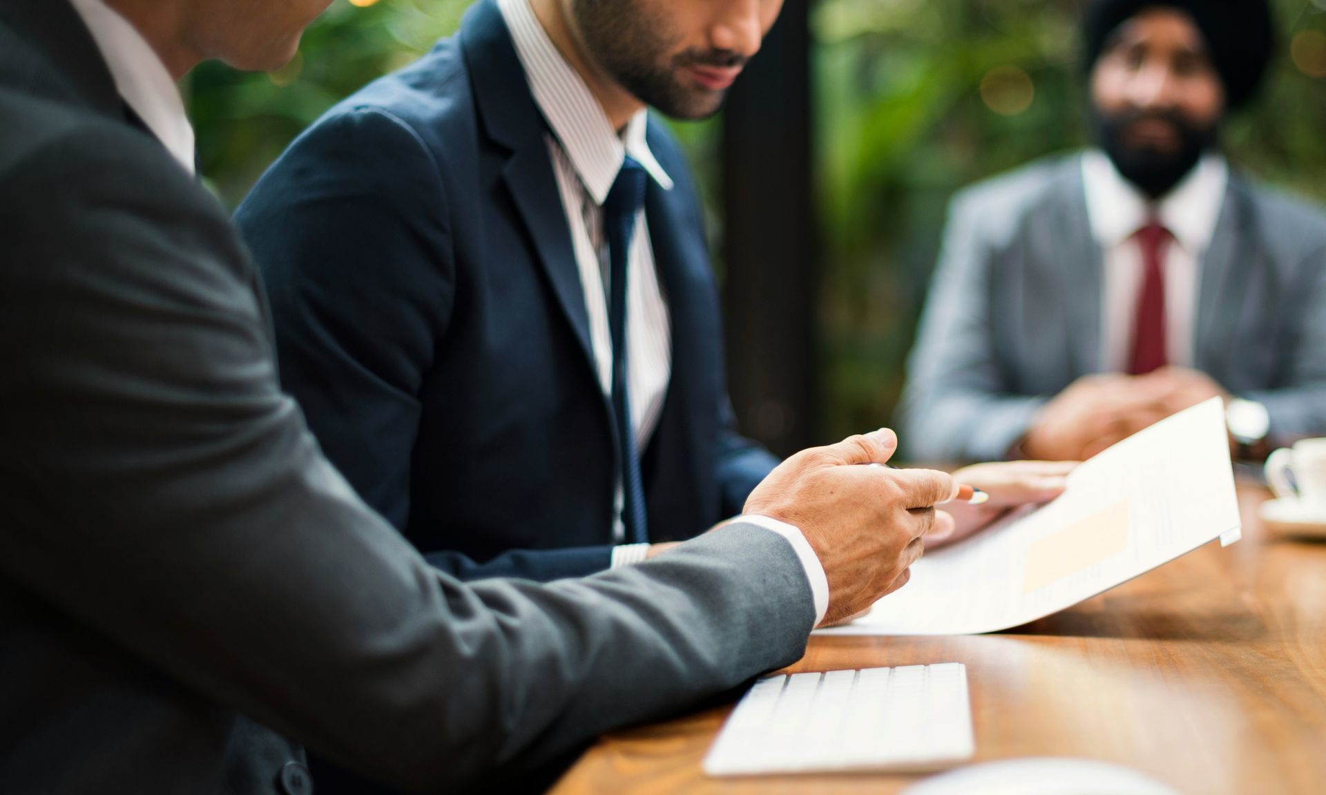 Three business professionals in suits review documents at a table outdoors.