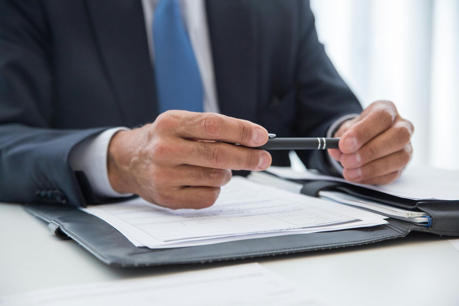 Person in suit holds a pen over documents in a binder; indoors.