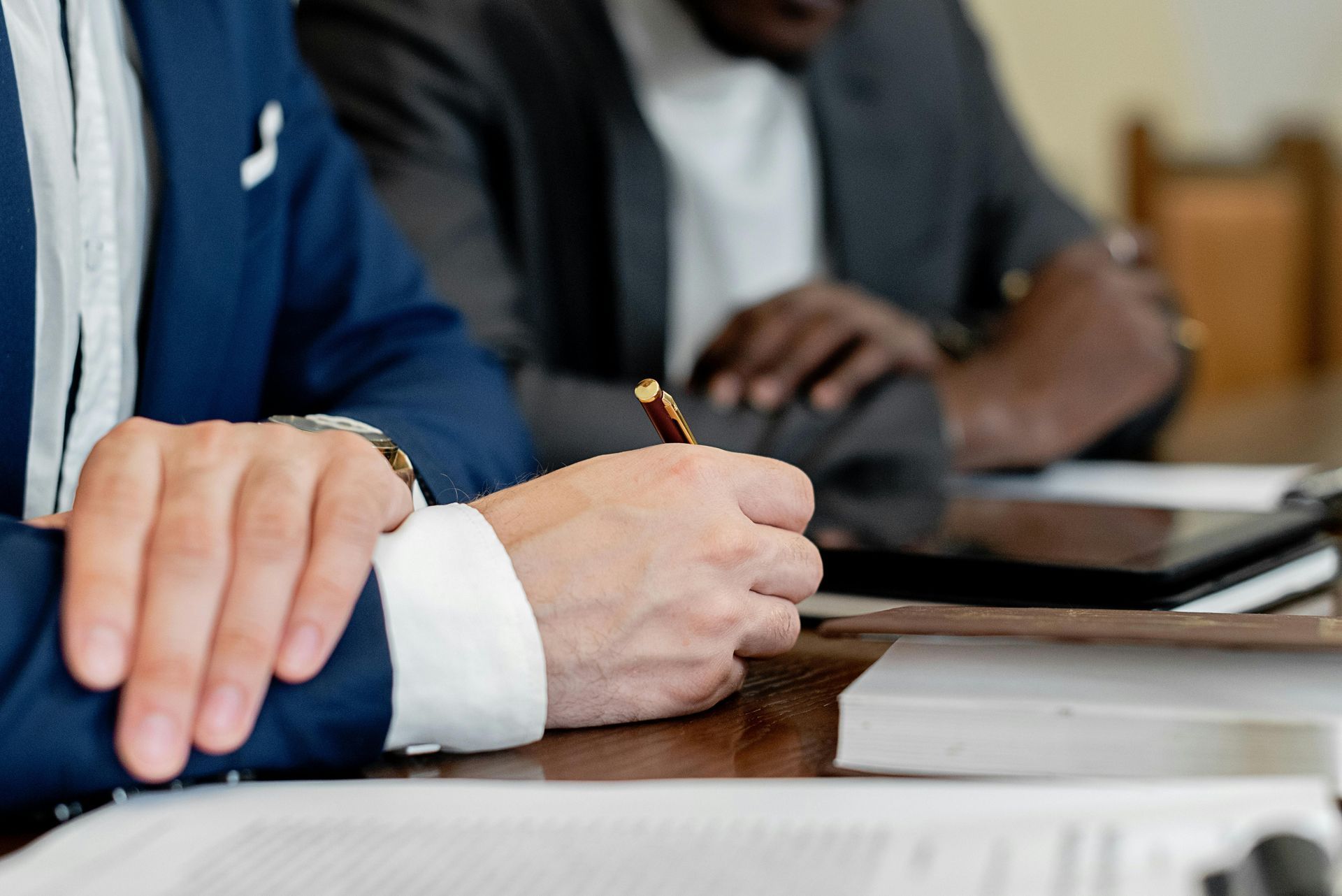 Person in blue suit writes with pen at a table. Another person, out of focus, is in background.