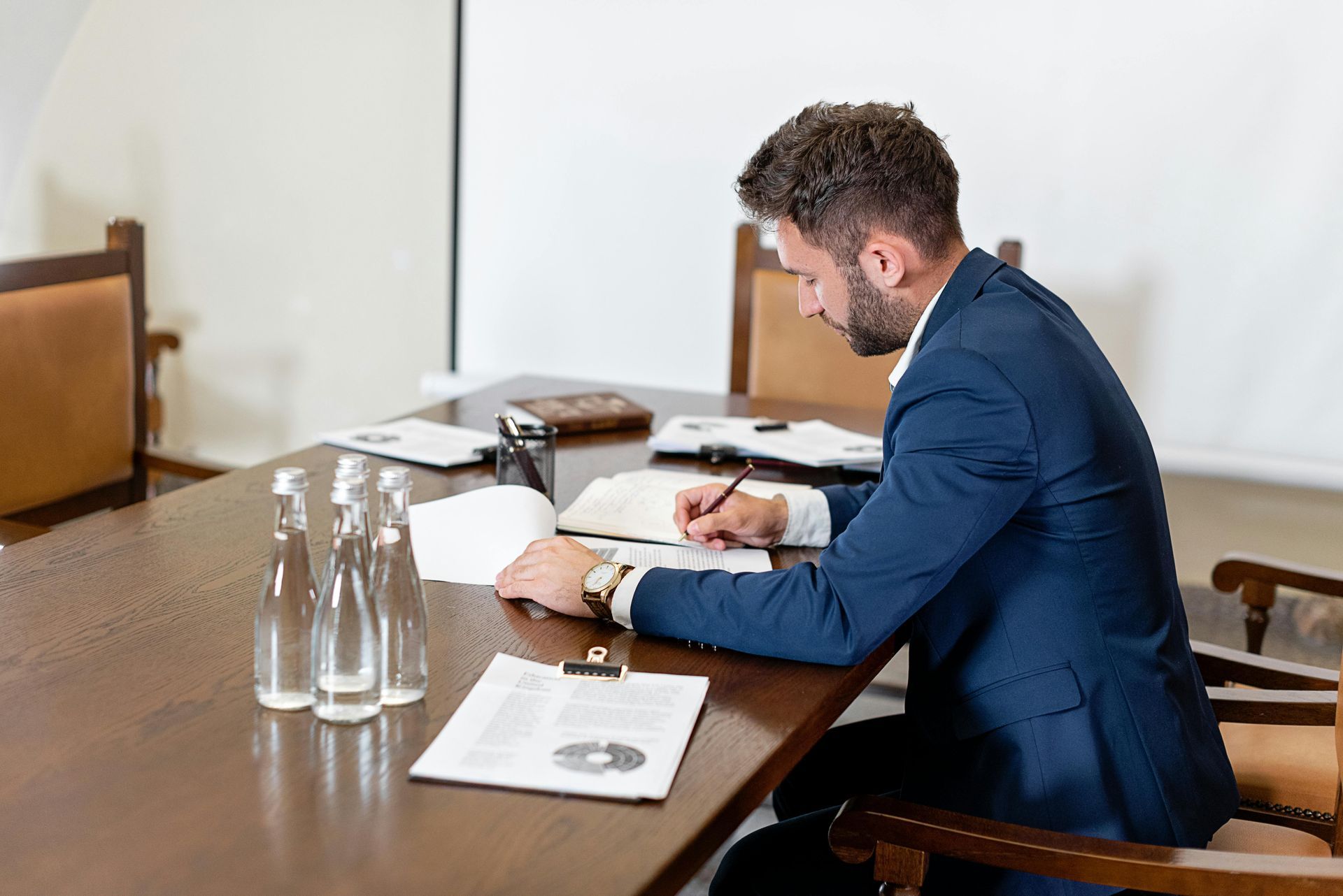 Man in a blue suit, writing at a conference table with papers and water bottles.