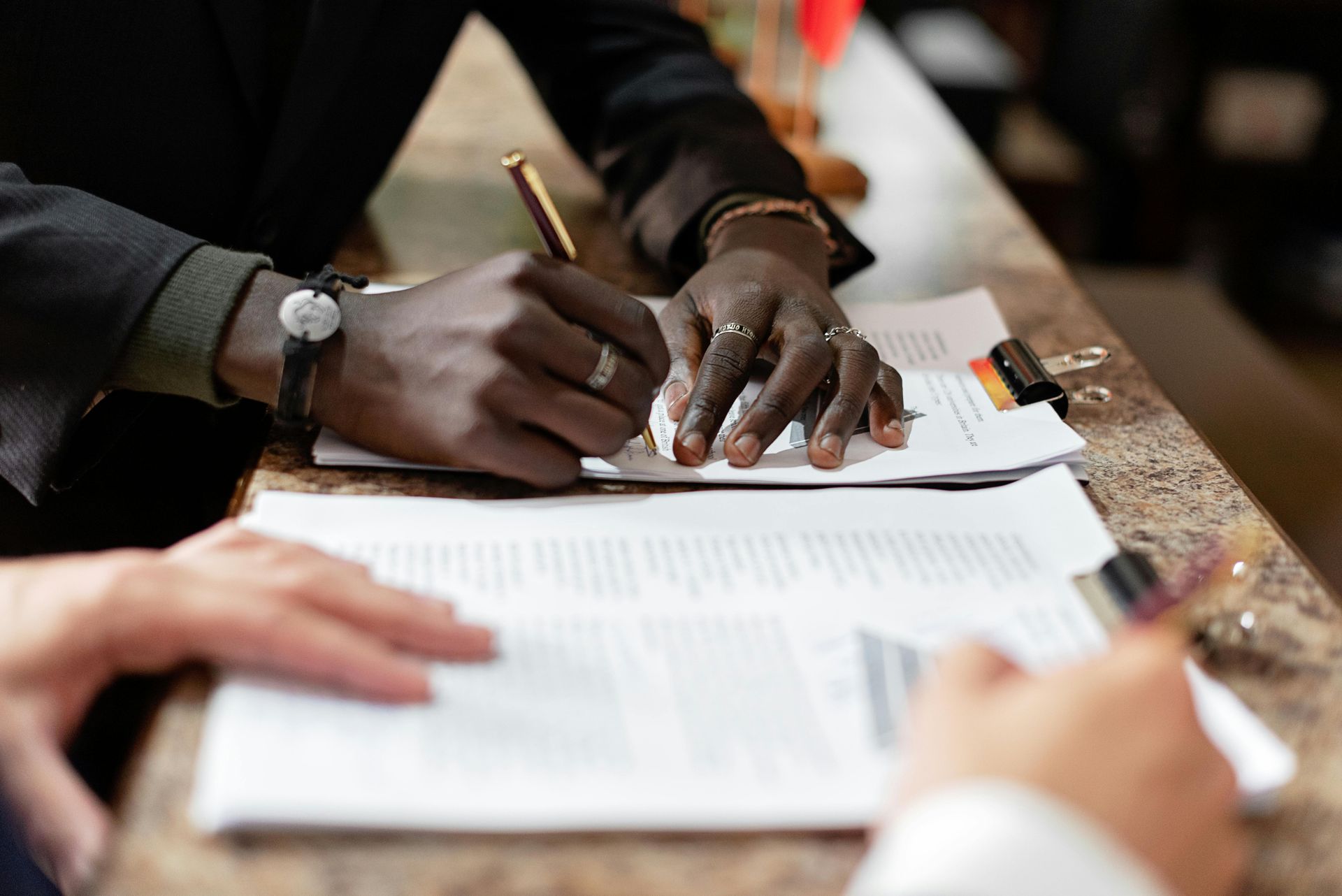 Person signing document, another person's hand in foreground. Brown counter, papers, pen, and hands.