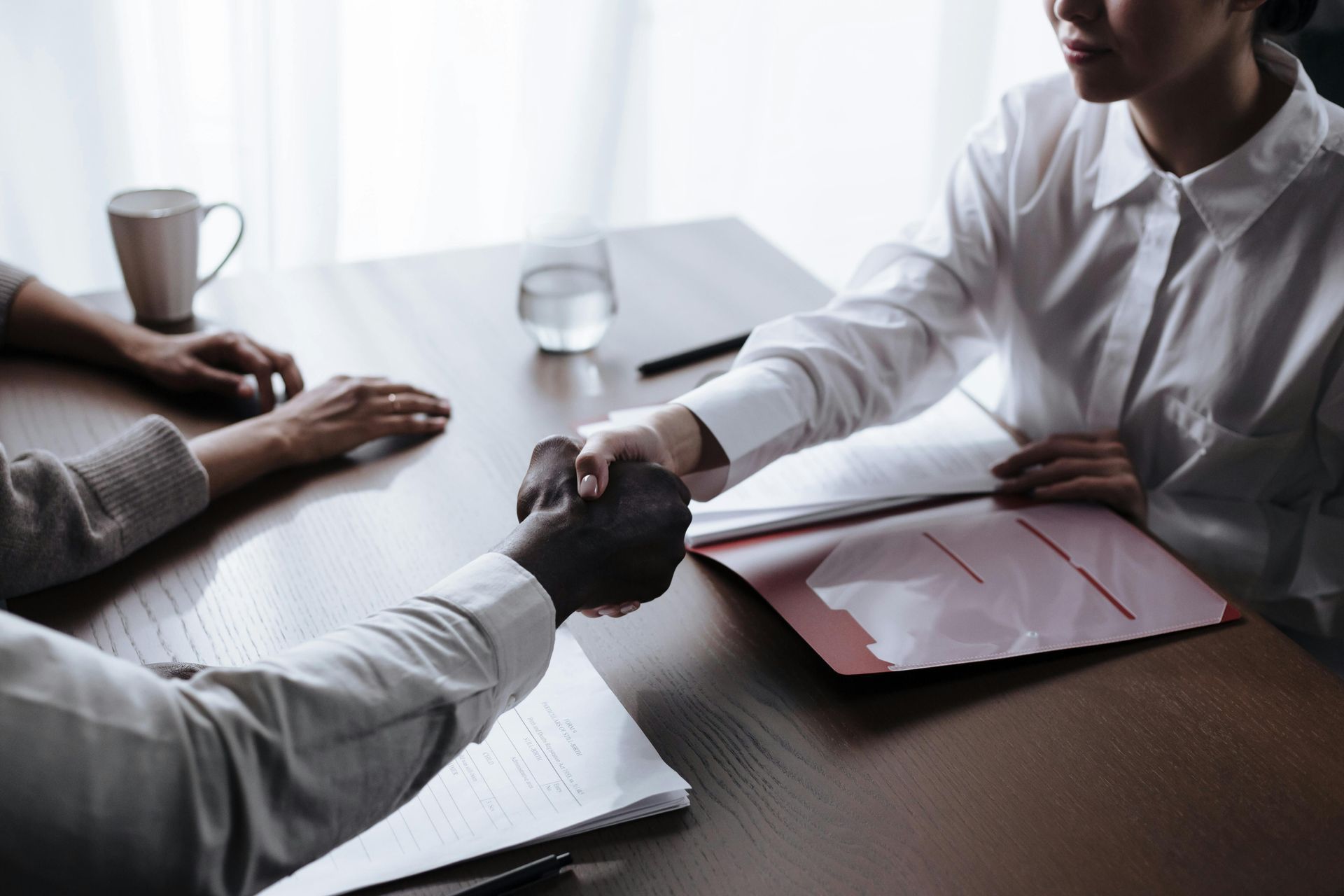 Two people shaking hands across a table; one is light-skinned, one dark-skinned, white shirt.