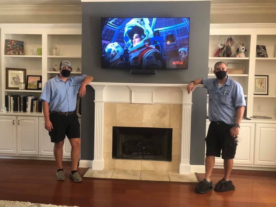 Two men in masks stand by a fireplace with a mounted TV. They are wearing shorts and leaning on the mantle.