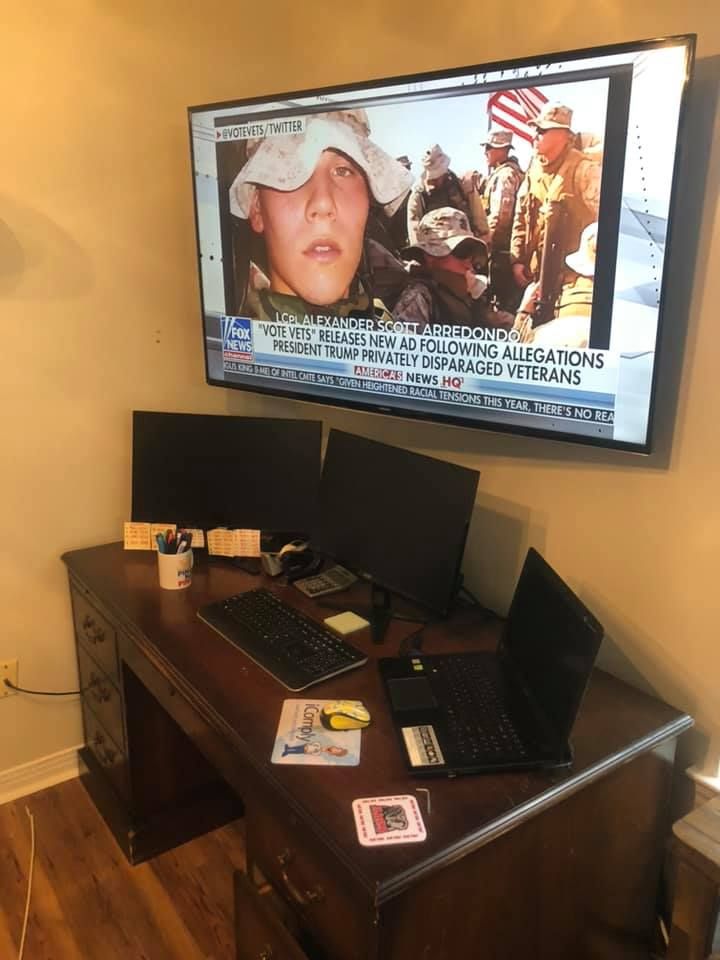A cluttered desk with two monitors, keyboard, and laptop, under a large TV showing a news broadcast.