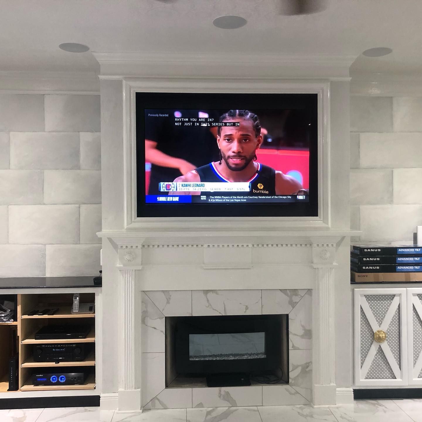 A living room with a television mounted above a fireplace featuring Kawhi Leonard on screen.