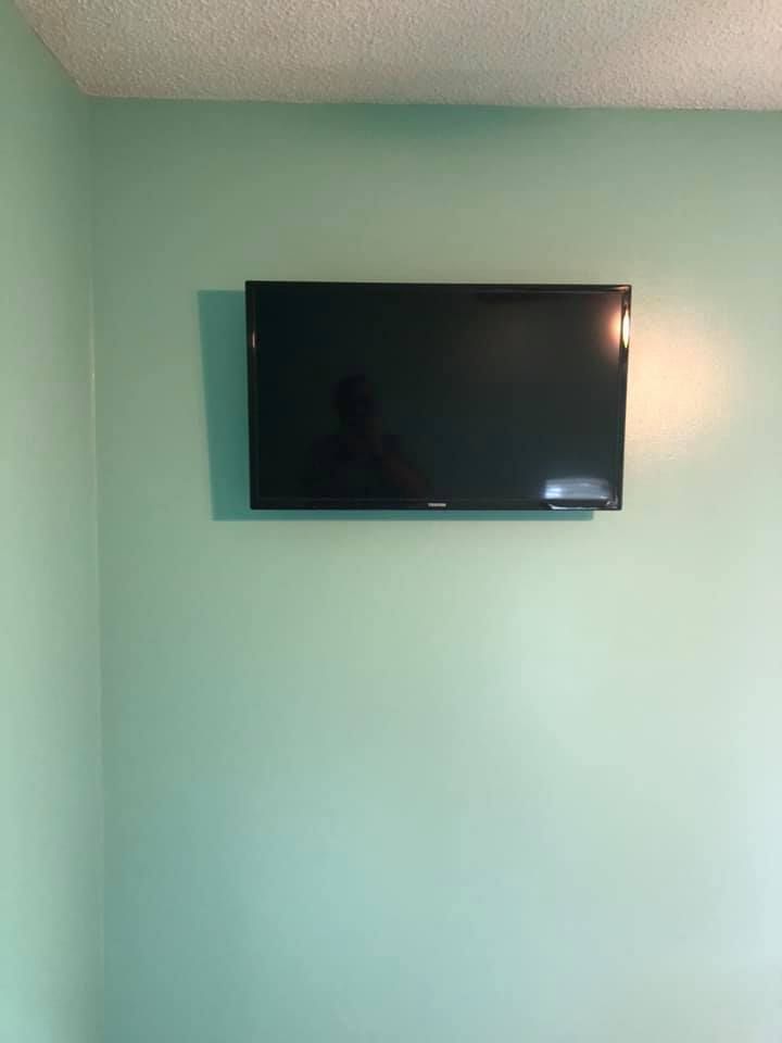 A flat-screen TV mounted on a light green wall. A person's reflection is visible on the dark screen.