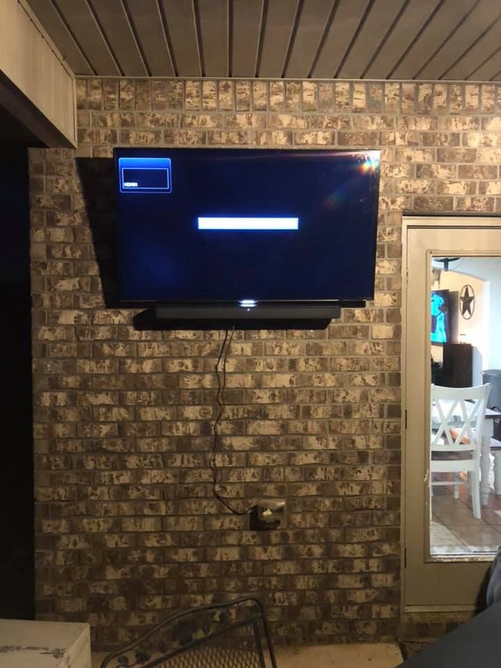 TV mounted on brick wall, loading screen displayed, soundbar below, cord hanging, outdoor setting.
