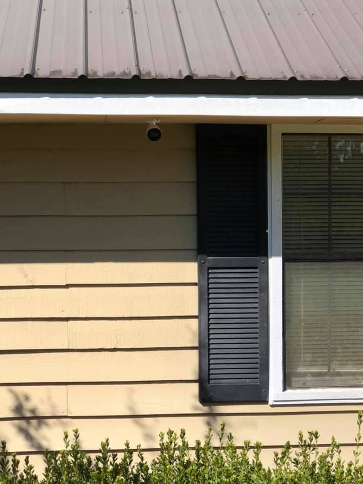 Exterior house wall with yellow siding, black shutter, window with blinds, and security camera. Brown metal roof.